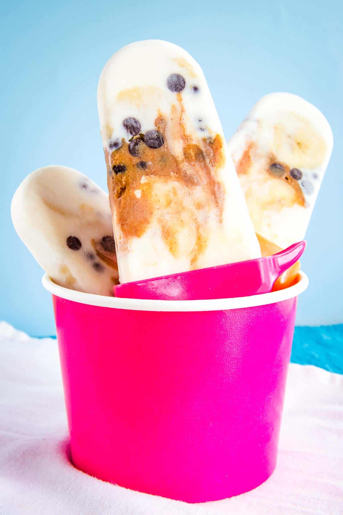 Three frozen yogurt popsicles with bananas, peanut butter, and chocolate chips in a pink cup.