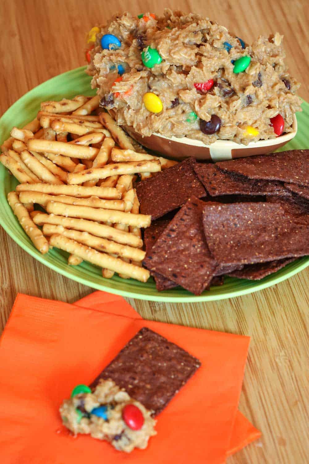 A grene plate with a bowl of cookie dough dip, pretzels, and chips.