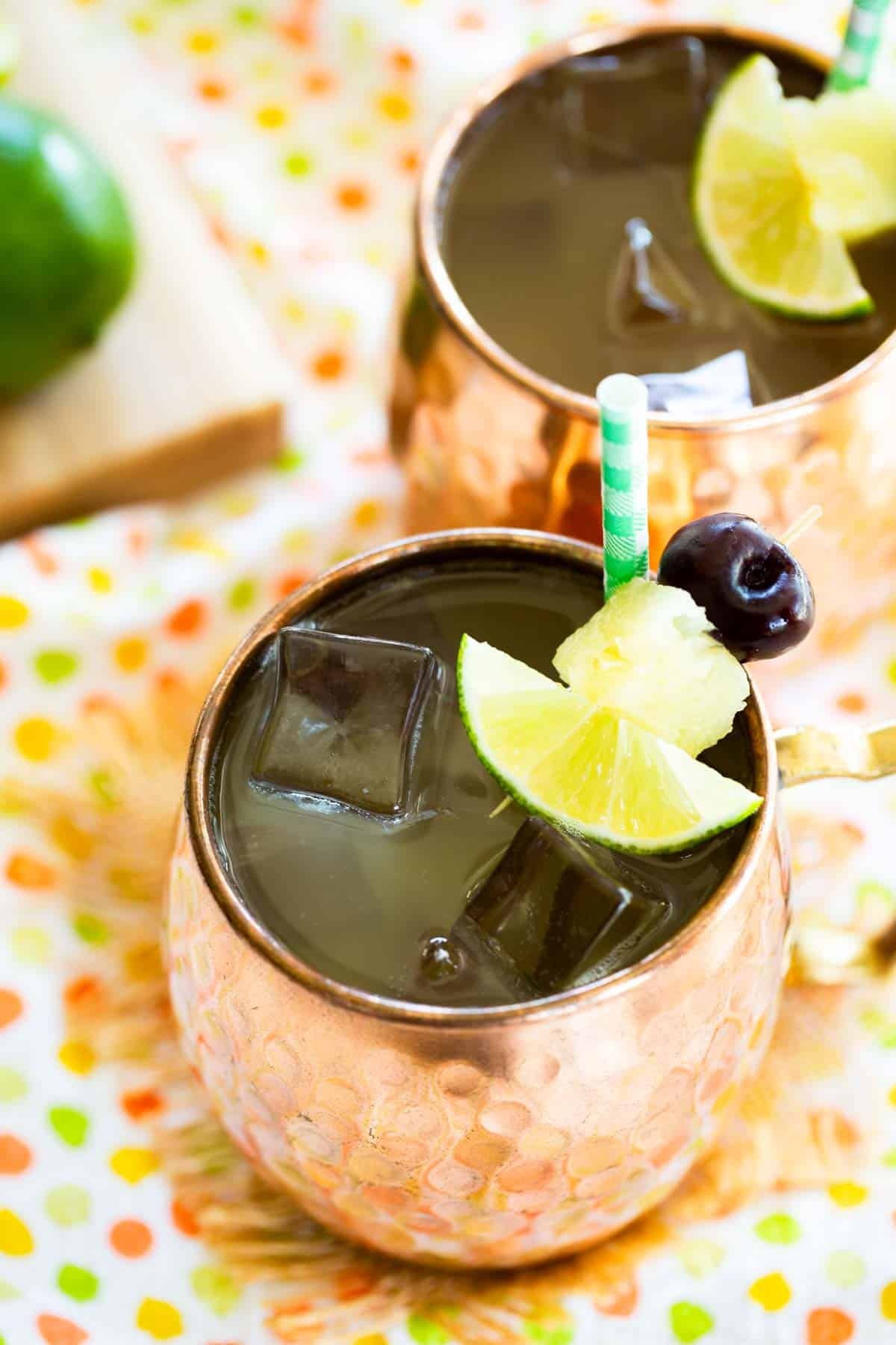Two copper mugs filled with a drink and garnished with a lime wedge, pineapple chunk, and cherry on a pick.