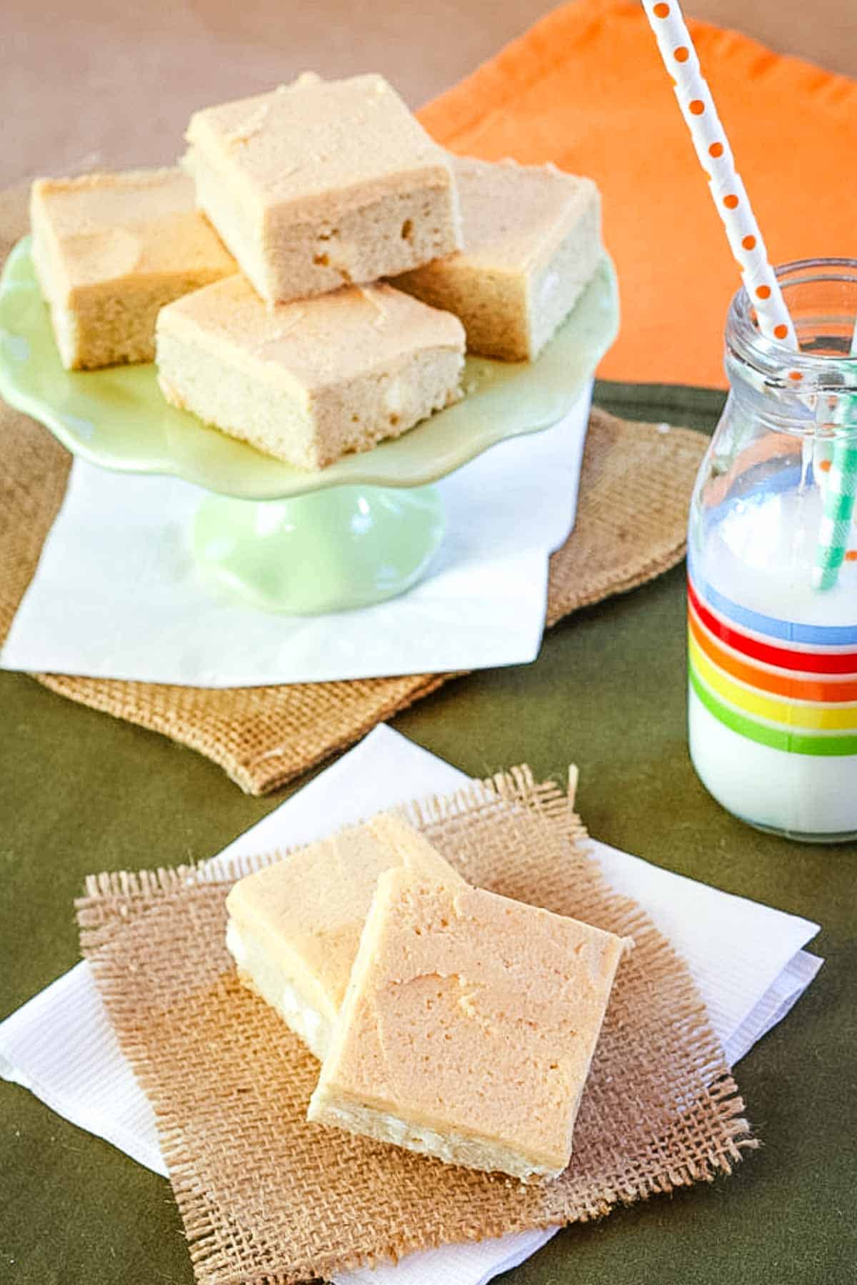 Frosted sugar cookie bars served next to a tall glass of milk and more cookie bars on a green cake stand.