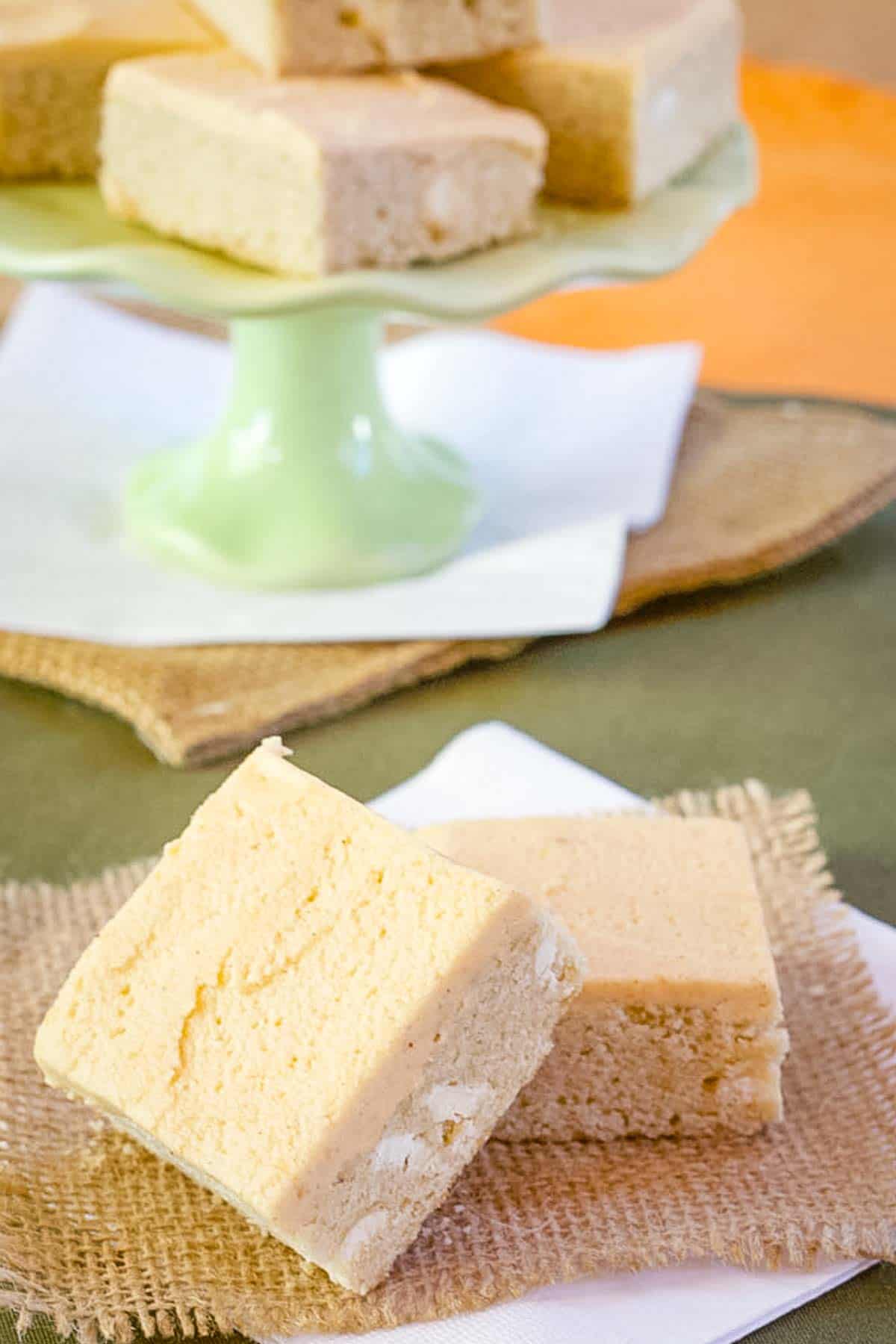 Two frosted sugar cookie bars in the foreground with a tray full of cookie bars in the background.