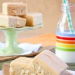 A cookie bar leaning against a second one on a napkin next to a glass of milk with more cookie bars on a cake stand in the background with text overlay that says "Pumpkin-Frosted Sugar Cookie Bars".