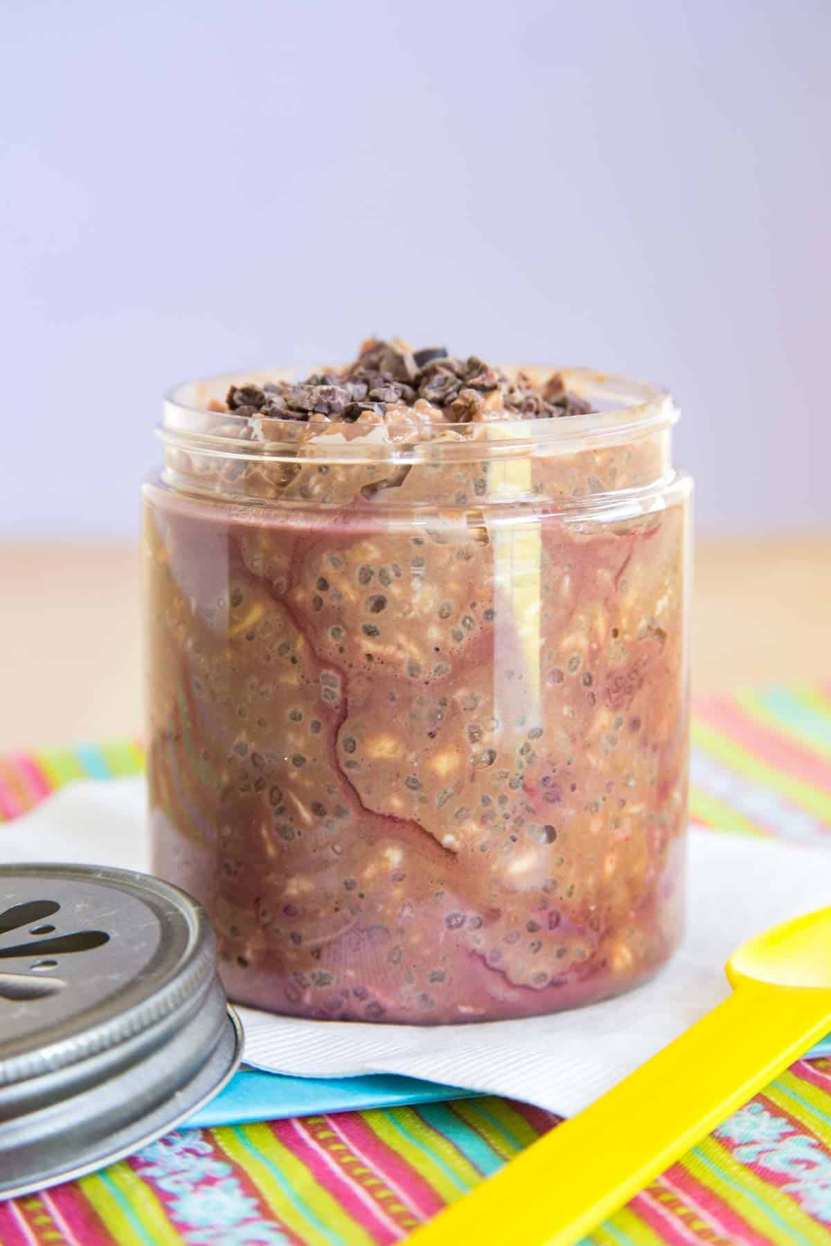 A jar of Raspberry Mocha Overnight Oats on top of a few colorful and white napkins with the lid and a plastic yellow spoon next to it.