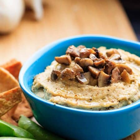 Mushroom hummus in a blue bowl with pita chips and sugar snap peas next to it.