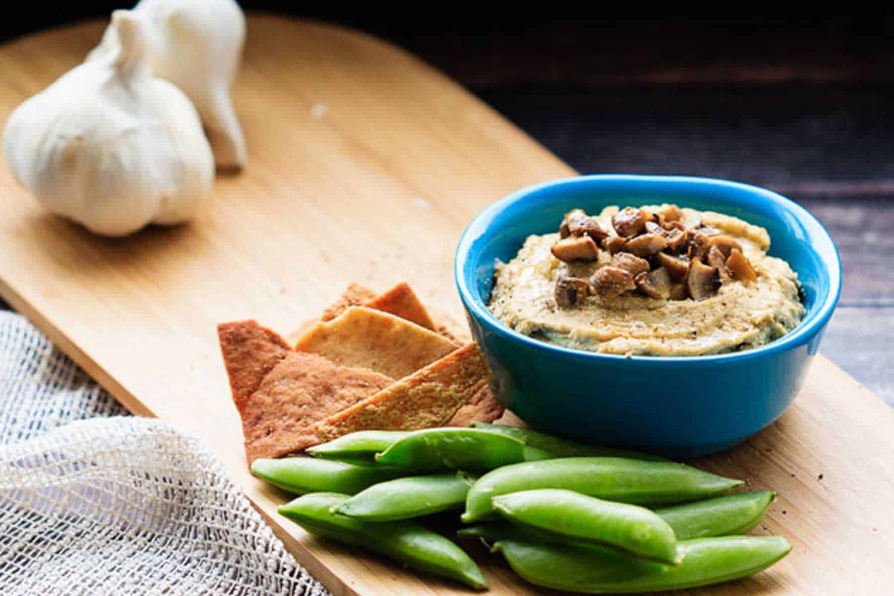 A blue bowl of hummus on a wooden board with sugar snap peas and pita chips plus heads of garlic in the background.