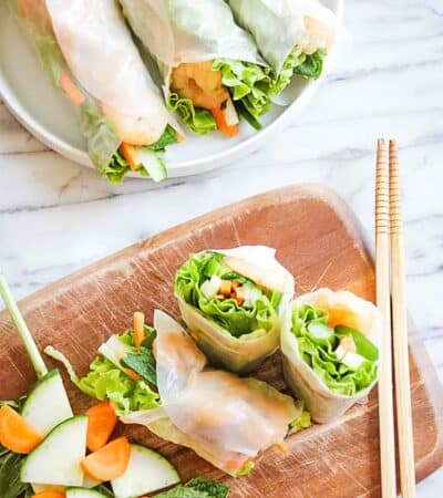 Rice paper rolls filled with shrimp and veggies on a cutting board with a set of chopsticks and more summer rolls on a plate.