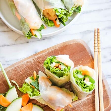 Rice paper rolls filled with shrimp and veggies on a cutting board with a set of chopsticks and more summer rolls on a plate.