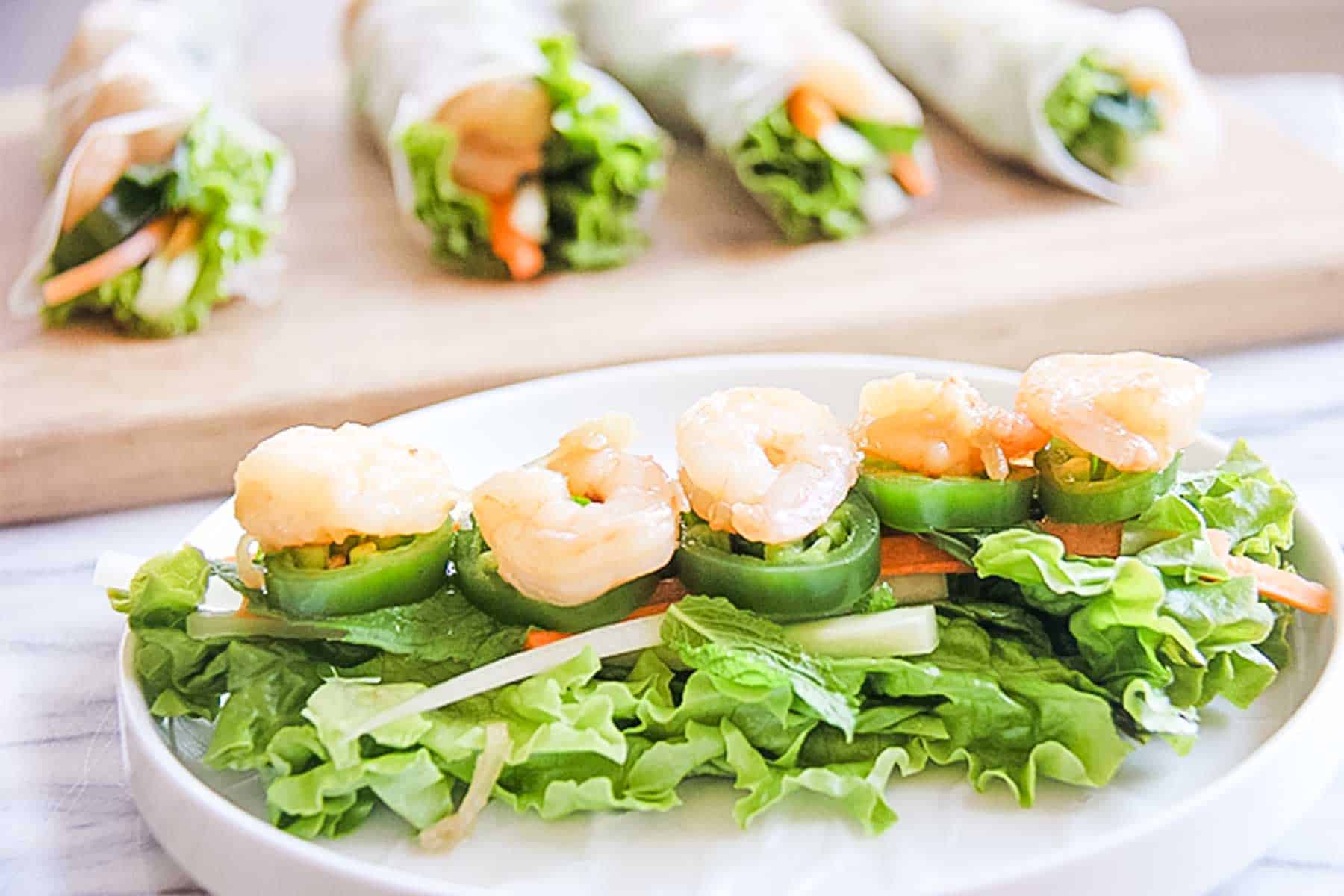 A bed of lettuce topped with julienned veggies, jalapeno slices and cooked shrimp on a white plate with fully made summer rolls on a cutting board in the background.