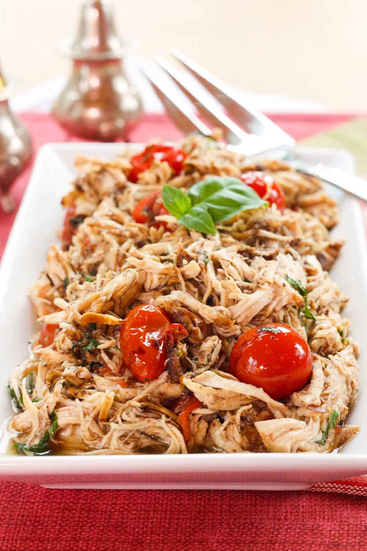 Slow Cooker Tomato Basil Chicken on a white rectangle serving platter with a meat fork resting on it.