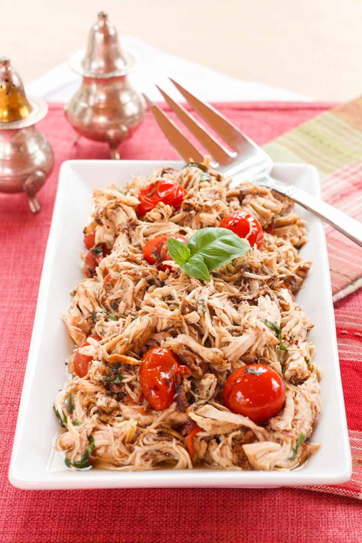 Shredded Balsamic Chicken with Tomatoes and a sprig of Basil on a white rectangular plate on top of a red placemat.