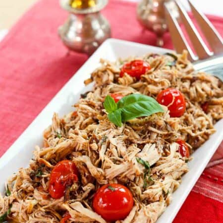 Shredded slow cooker chicken with tomatoes and basil on a white plate on top of a red placemat with silver salt and pepper shakers.