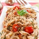 Shredded chicken with tomatoes on a rectangular white plate with text overlay that says "Slow Cooker Tomato Basil Chicken".