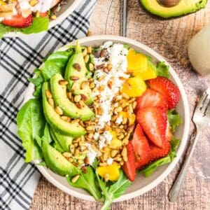 A bowl of salad with strawberries, avocado, and mango with pine nuts and goat cheese.