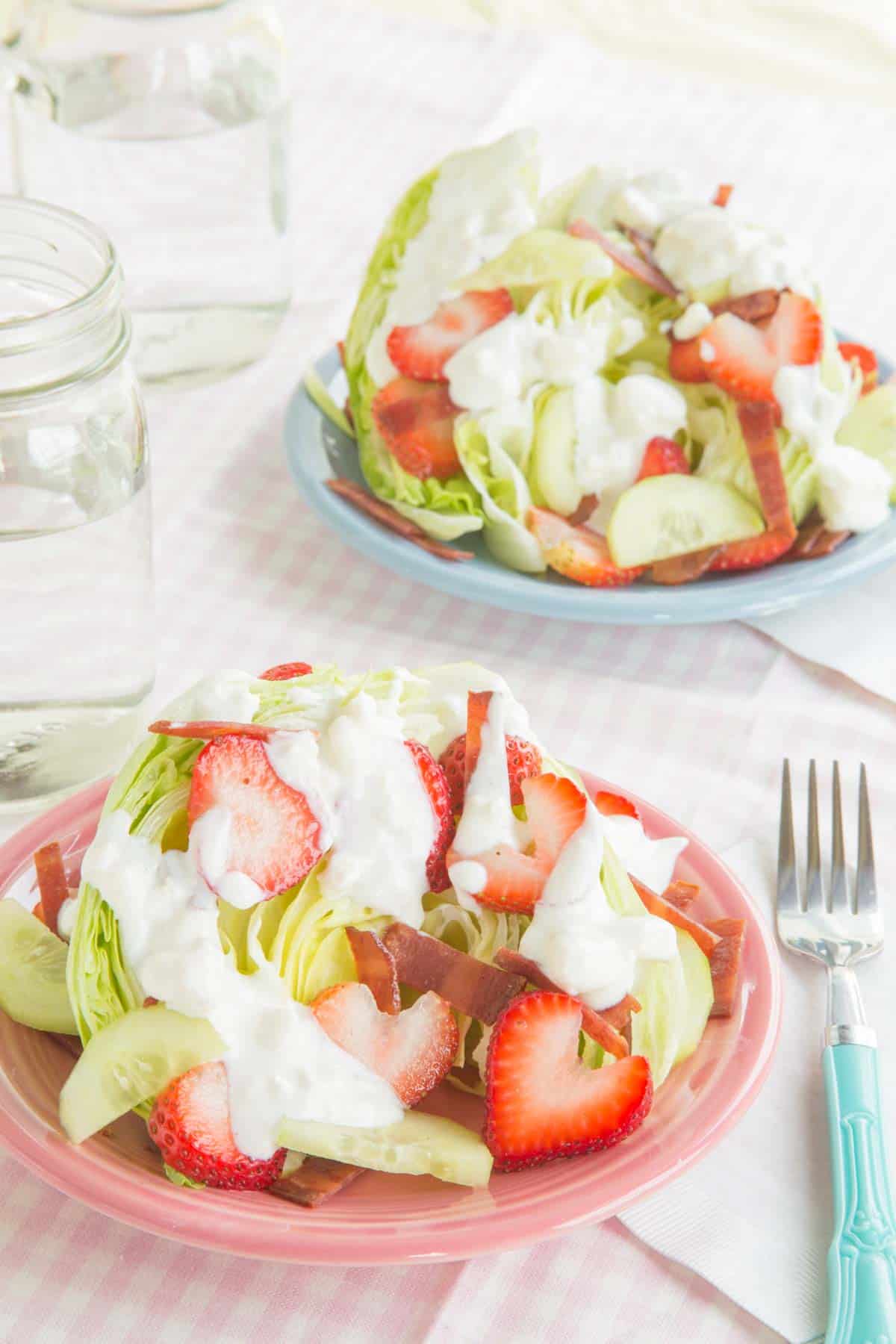 Two plates of an iceberg lettuce wedge topped with bacon, sliced strawberries and cucumbers, and blue cheese dressing.