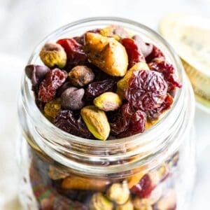 Looking down into a jar of snack mix with pistachios, dried cherries, chocolate chips, and pretzel nuggets.