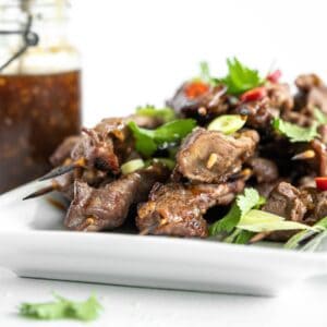 grilled teriyaki beef sticks on a white platter garnished with cilantro and scallions.