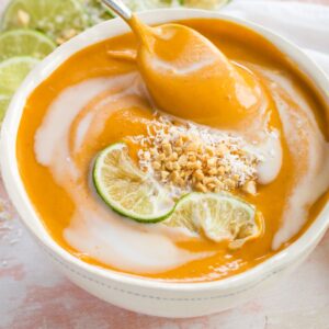 A spoon in a bowl of curried butternut squash soup swirled with coconut milk and garnished with choppe dpeanuts, shredded coconut, and lime.