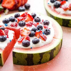 A slice being pulled away from a watermelon pizza topped with whipped cream and berries.