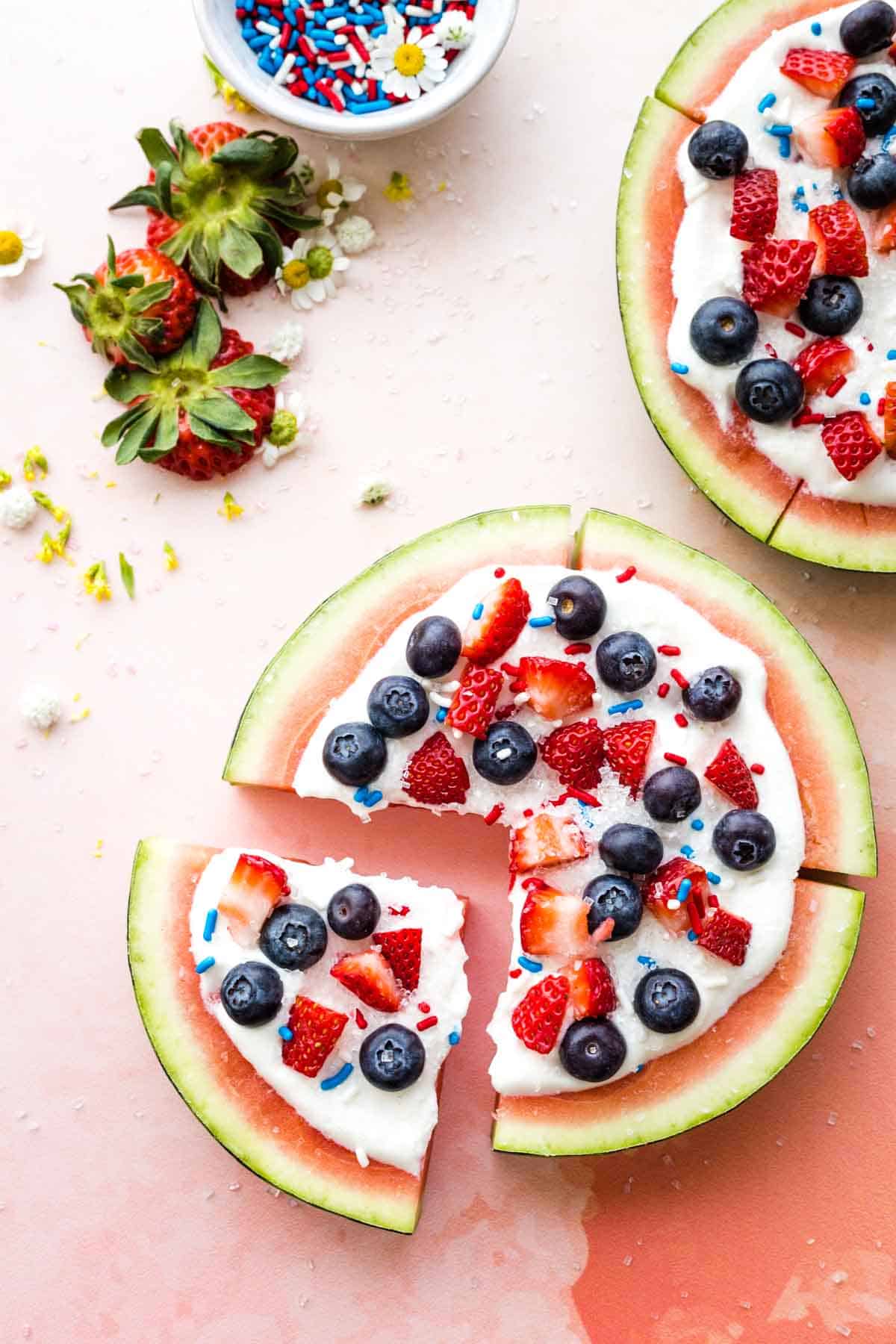 A birdseye view of a round of watermelon topped with berries, whipped cream, and sprinkled, and cut into four wedges.