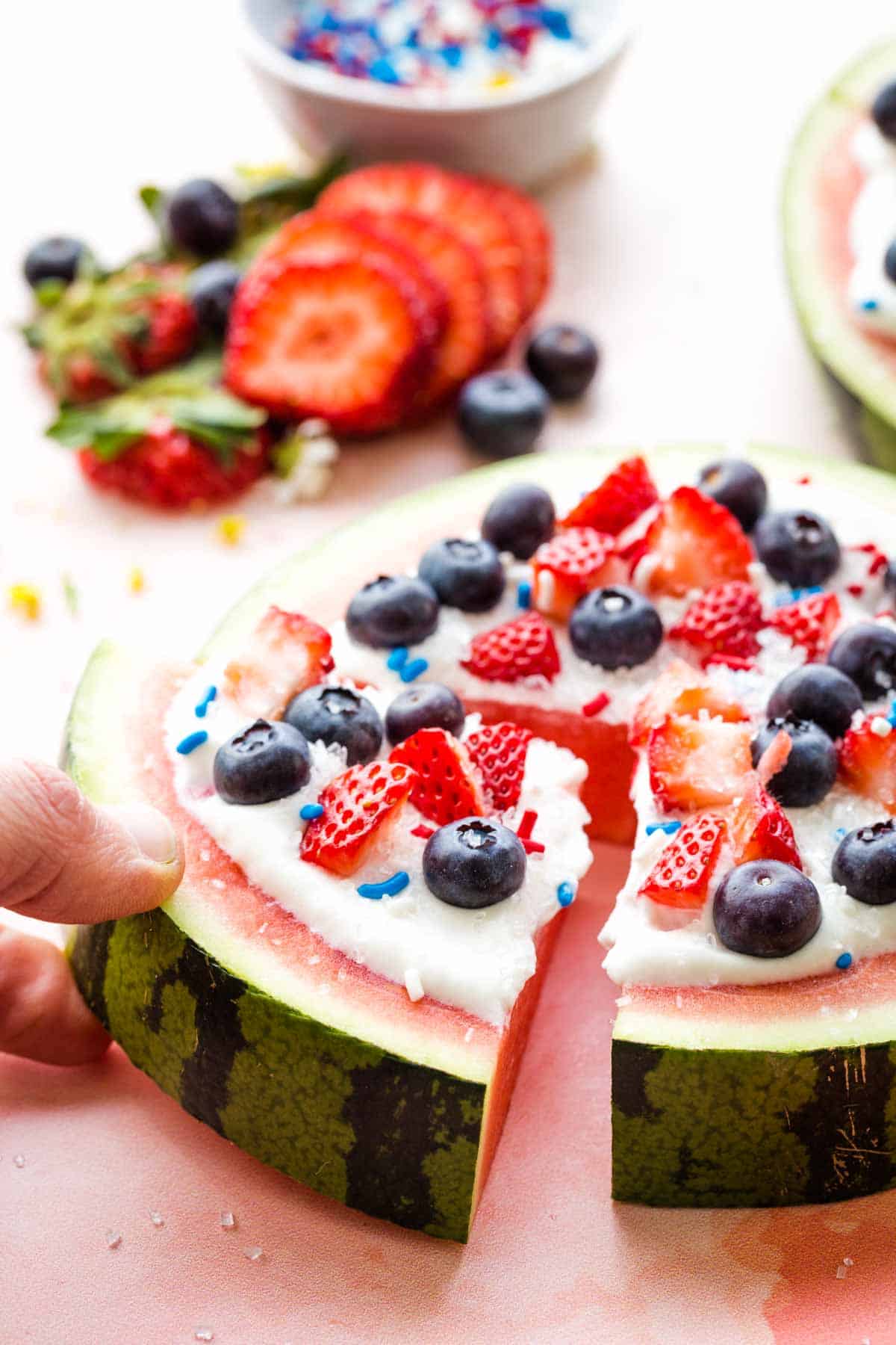 A hand picking up a slice of watermelon fruit pizza with berries and whipped cream.