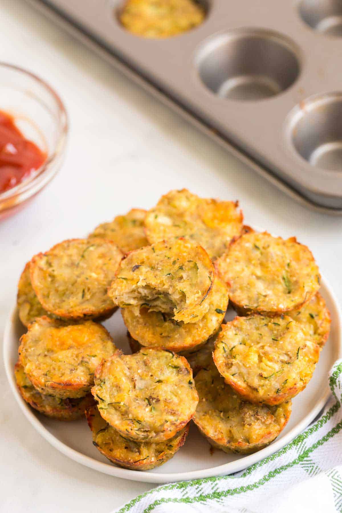 A plate of zucchini tots next to a bowl of ketchup and a muffin tin.