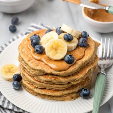 A stack of gluten-free protein pancakes topped with blueberries and sliced bananas with a fork alongside.
