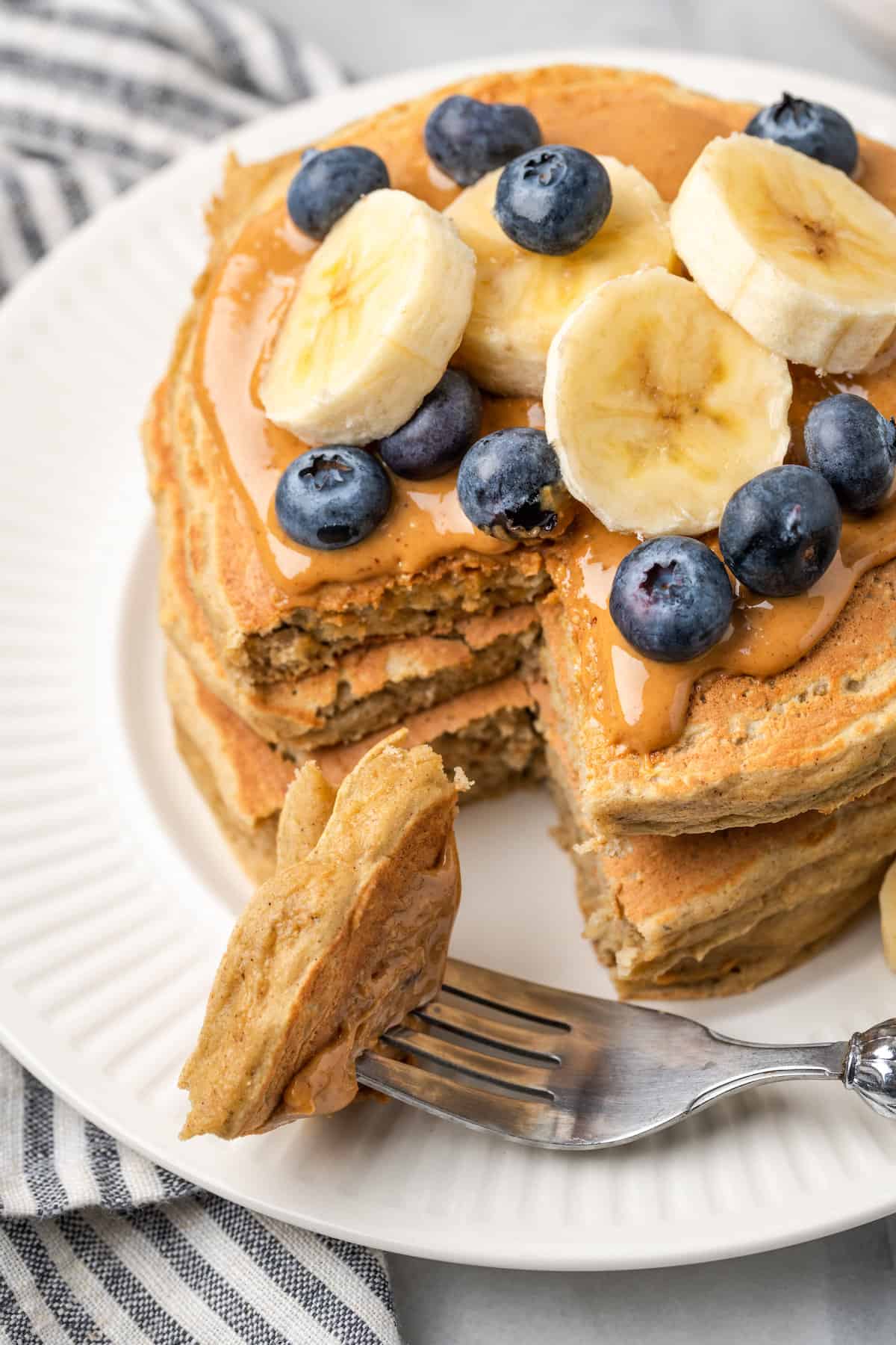 A fork cuts into a stack of gluten-free protein pancakes topped with blueberries and sliced bananas.