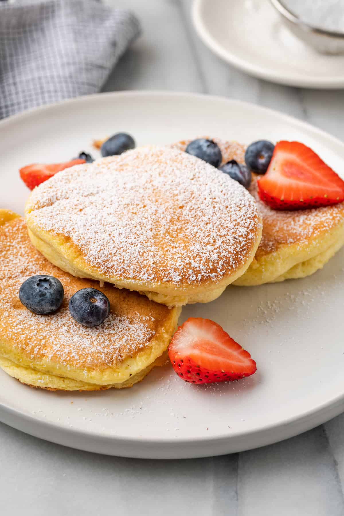 Gluten-free souffle pancakes on a plate, and dusted with powdered sugar and topped with strawberries and blueberries.