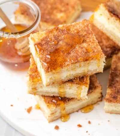 A stack of sopapilla cheesecake bars is shown on a plate drizzled with honey.