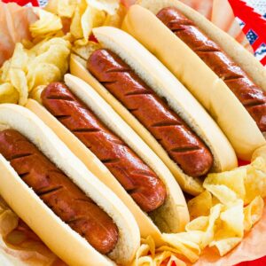 Four hot dogs in buns lined up in a basket surrounded by potato chips.