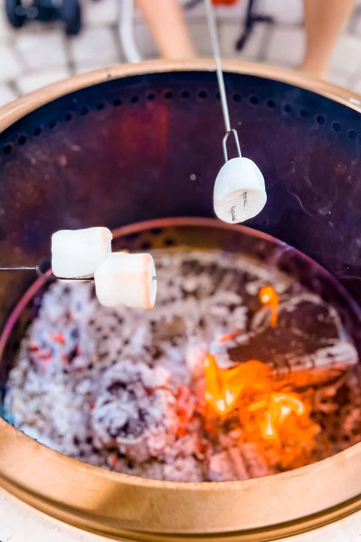 Three marshmallows being held on skewers and toasting over a fire pit.