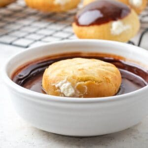 A round filled donut floating upside down in a bowl of chocolate glaze in front of a rack with more donuts on it.
