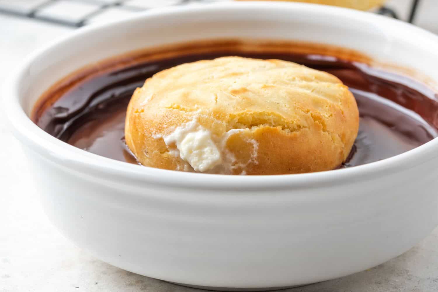 A round filled donut floating upside down in a bowl of chocolate glaze.