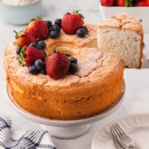 An angel food cake topped with berries one a cake pedestal with one slice being lifted out on a spatula.