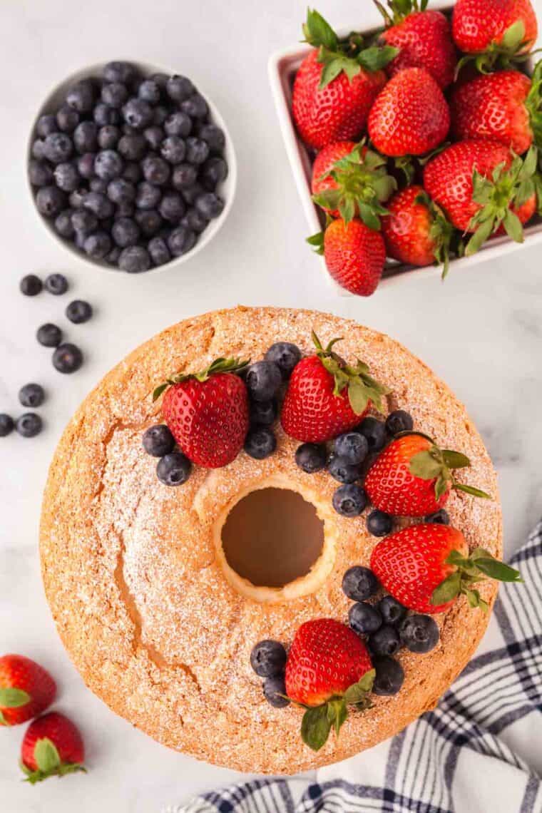 An overhead photo of gluten-free angel food cake topped with blueberries and strawberries, with more berries in the background.