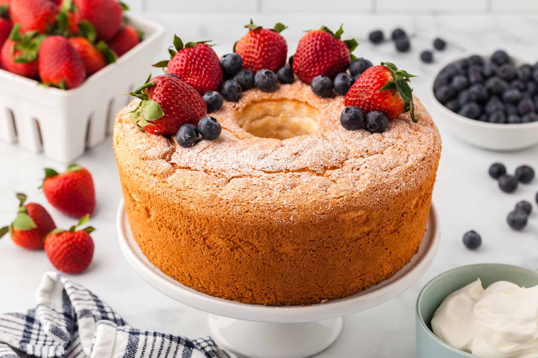 A whole gluten-free angel food cake is shown on a cake stand topped with strawberries and blueberries.