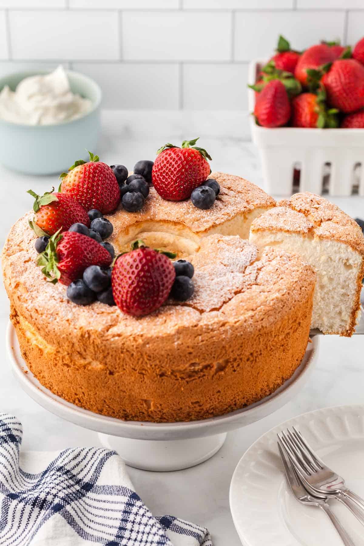 A gluten-free angel food cake is shown on a cake stand topped with strawberries and blueberries with a slice cut out.