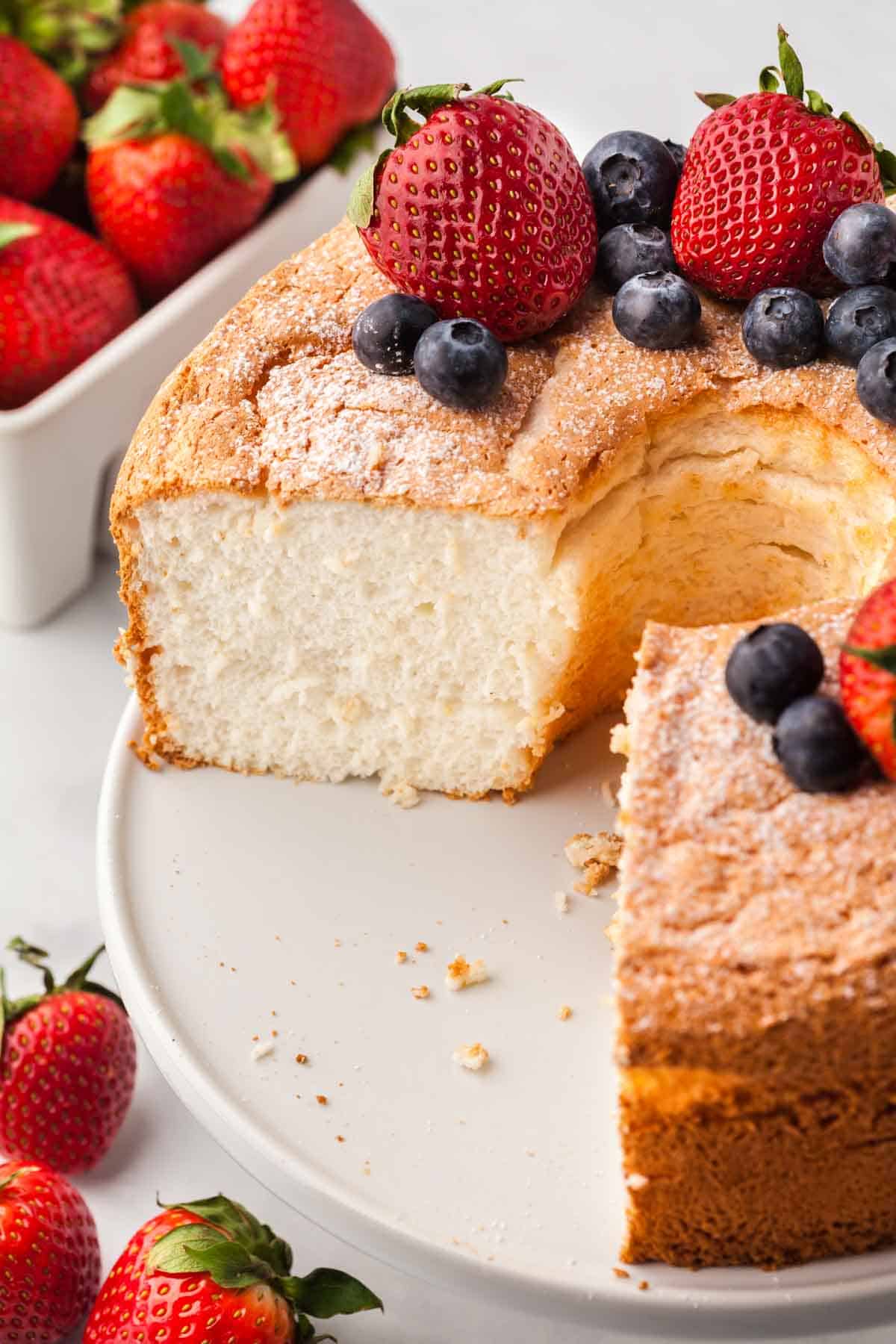 A gluten-free angel food cake is shown on a cake stand topped with strawberries and blueberries.