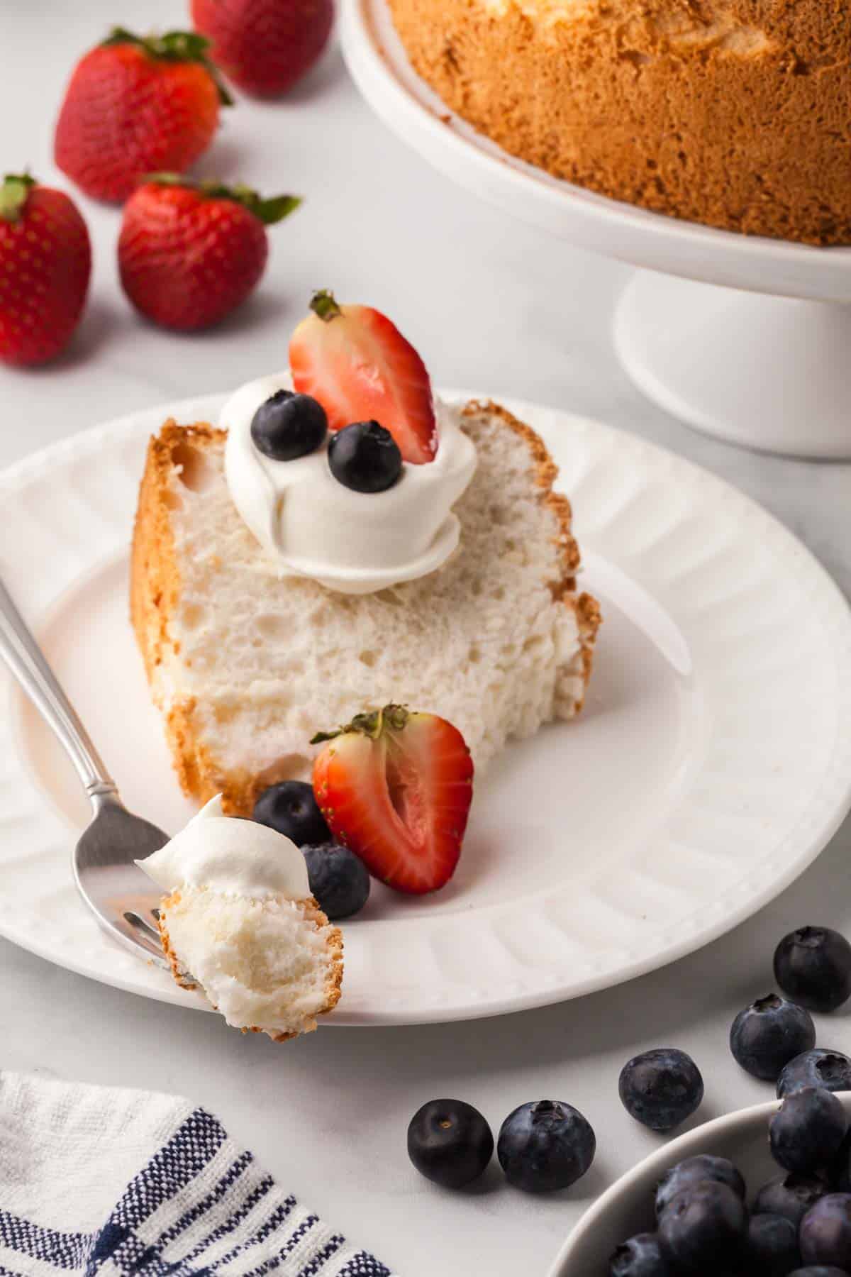 A slice of gluten-free angel food cake is shown on a white plate topped with whipped cream and berries, with a fork alongside.