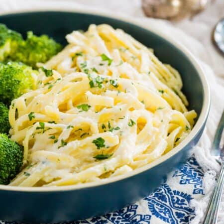 A serving of gluten free fettuccine alfredo garnished with chopped parsley in a blue dish with a side of broccoli.