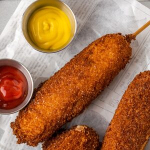 Three golden fried Korean corn dogs on wax paper with small dishes of ketchup and mustard.