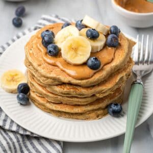 A stack of gluten free protein pancakes on a plate topped with peanut butter, blueberries, and banana slices with a fork resting on the plate next to them.