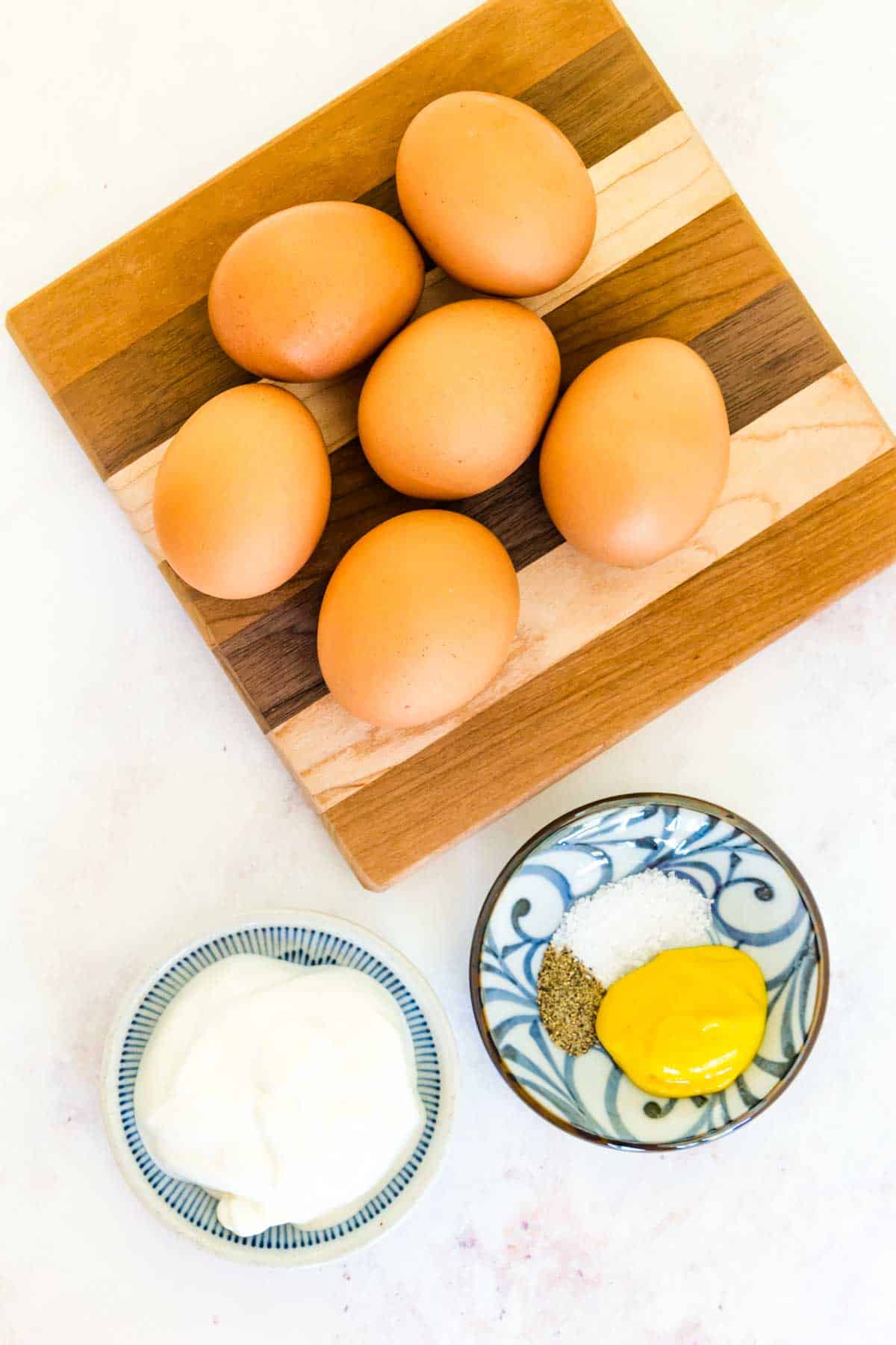 Six eggs on a wooden cutting board with small bowls of Greek yogurt, mustard, salt, and pepper.