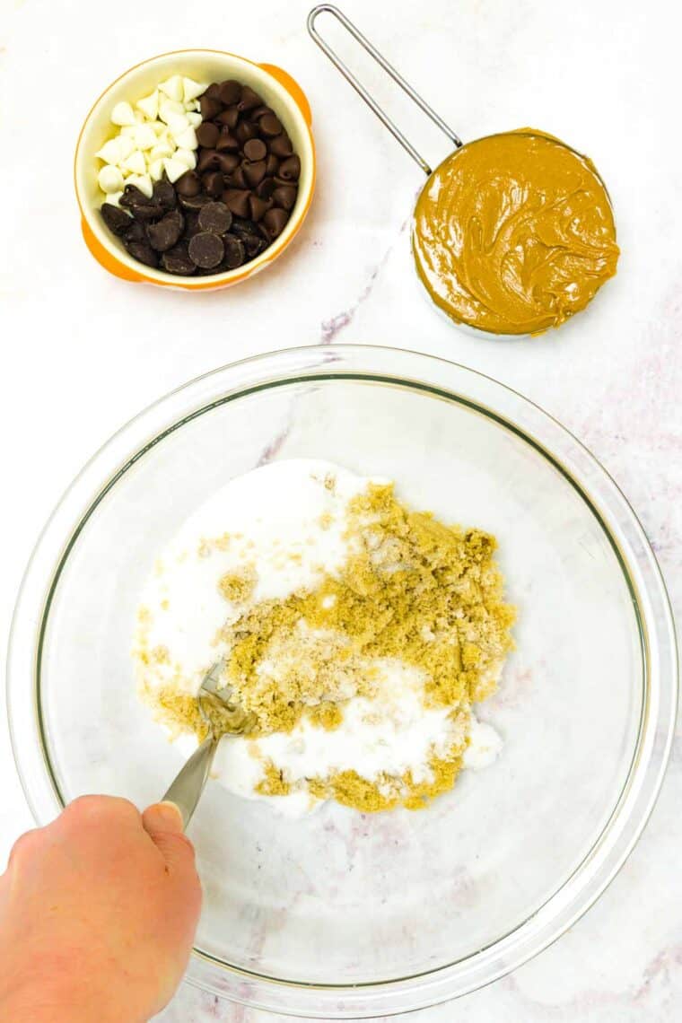 White and brown sugar being mixed in a glass bowl with a fork with a measuring cup of peanut butter and a bowl of chocolate chips next to it.