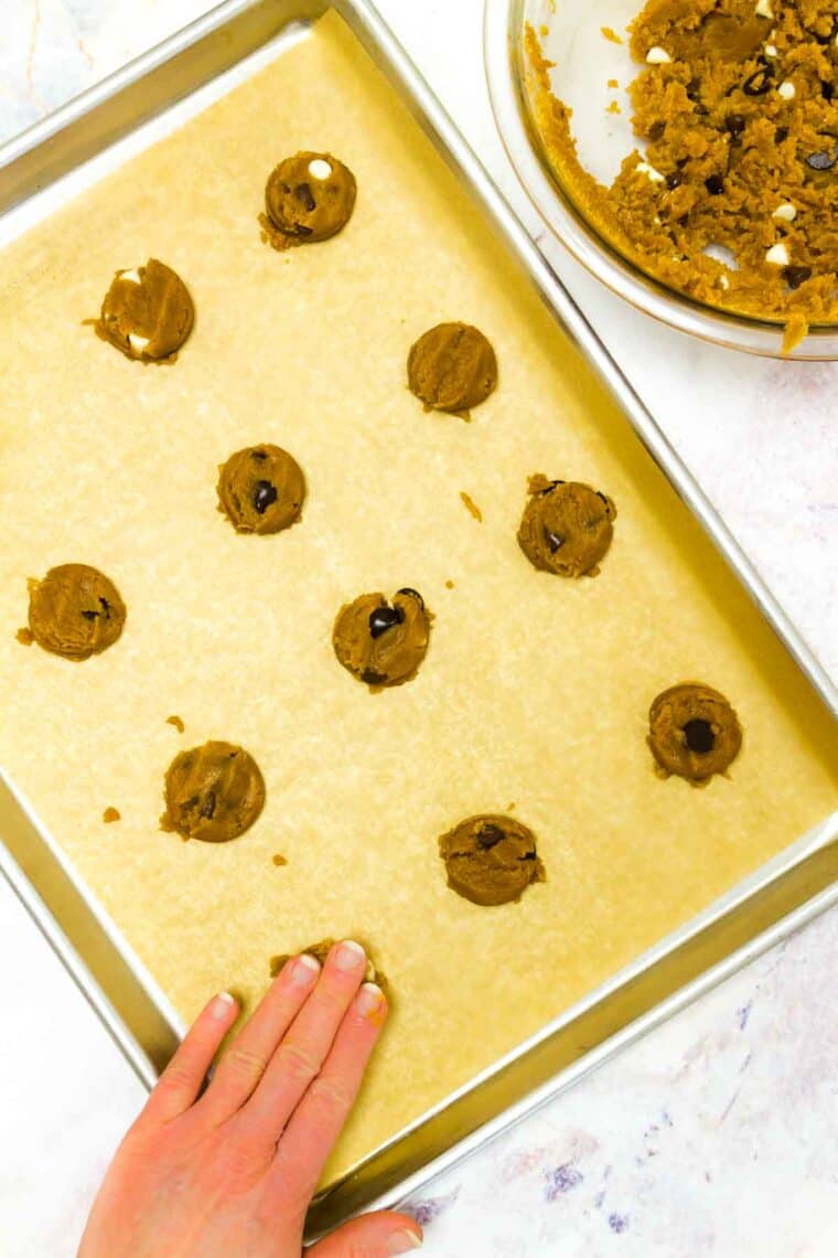 A hand patting down on of the balls of cookie dough on a parchment-lined sheet pan.