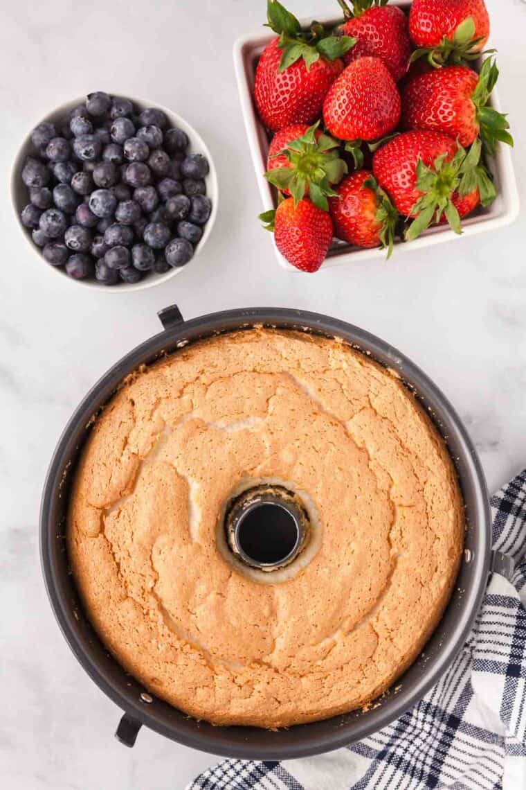 An overhead photo of a gluten-free angel food cake in a tube pan with a bowl of blueberries and a container of strawberries.