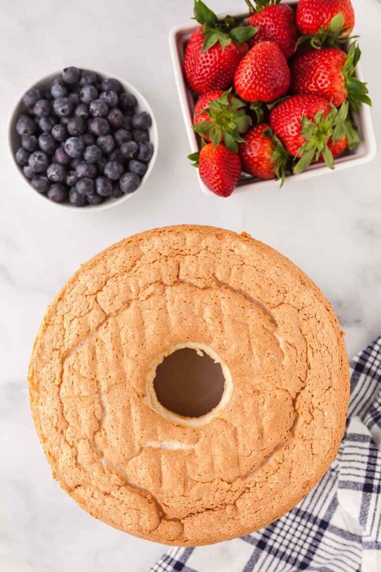 An overhead photo of a gluten-free angel food cake on a rack with a bowl of blueberries and a container of strawberries.