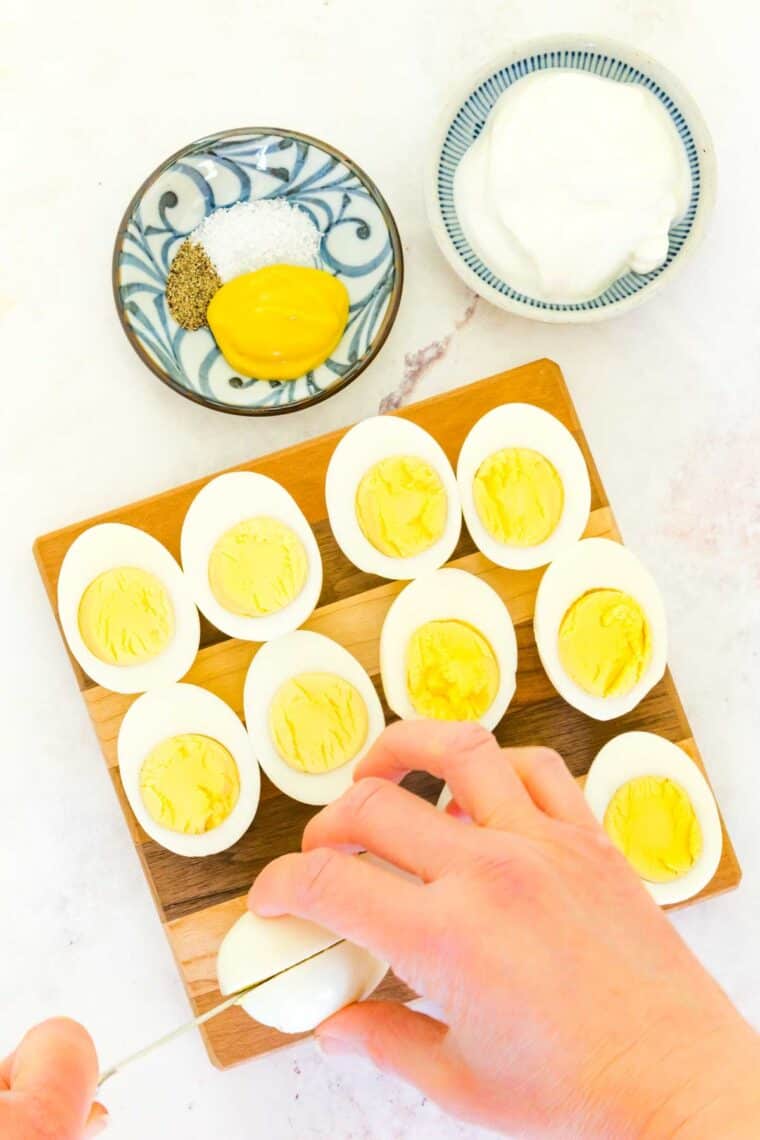 Hard boiled eggs cut in half on a cutting board with one being cut with small bowls of Greek yogurt, mustard, salt, and pepper.