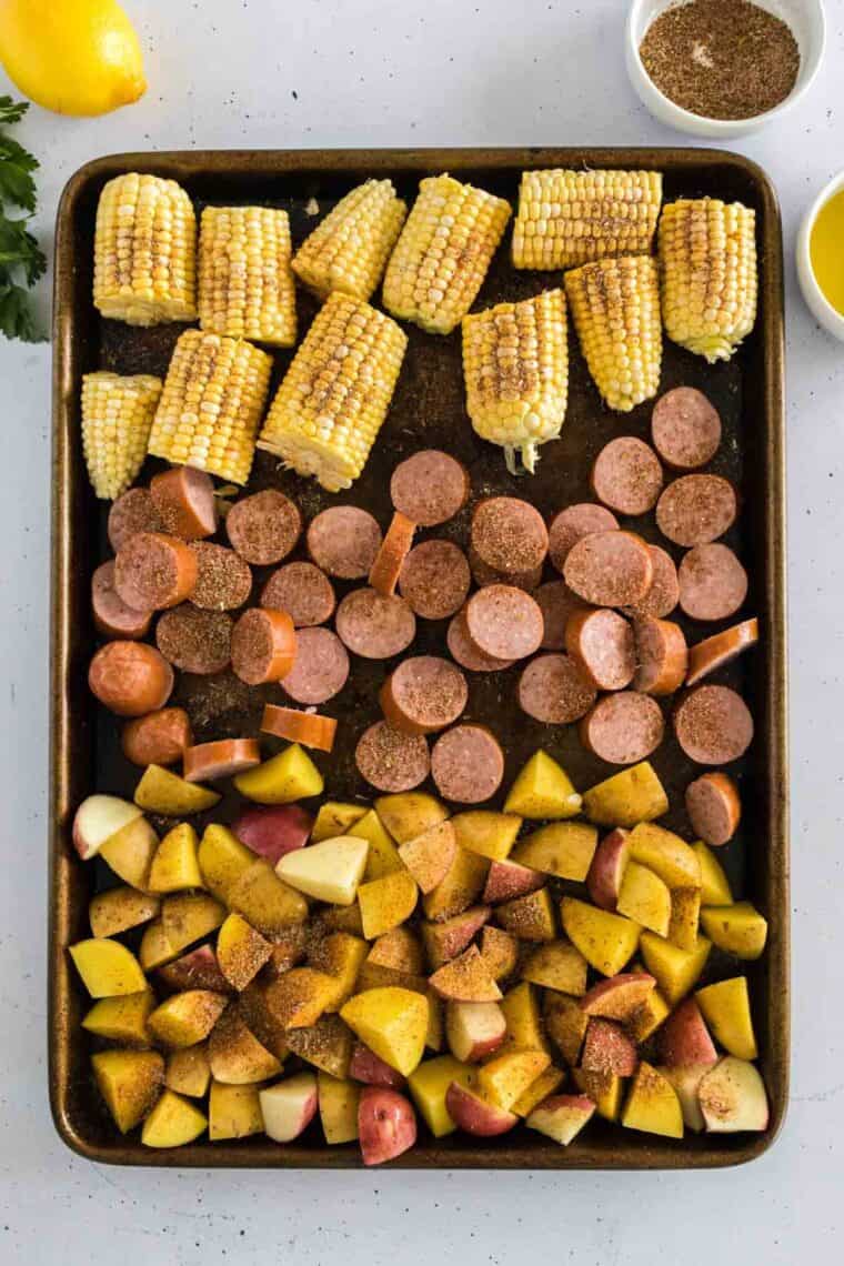 Corn on the cob, sausage, and cut potatoes are arranged on sheet pan.