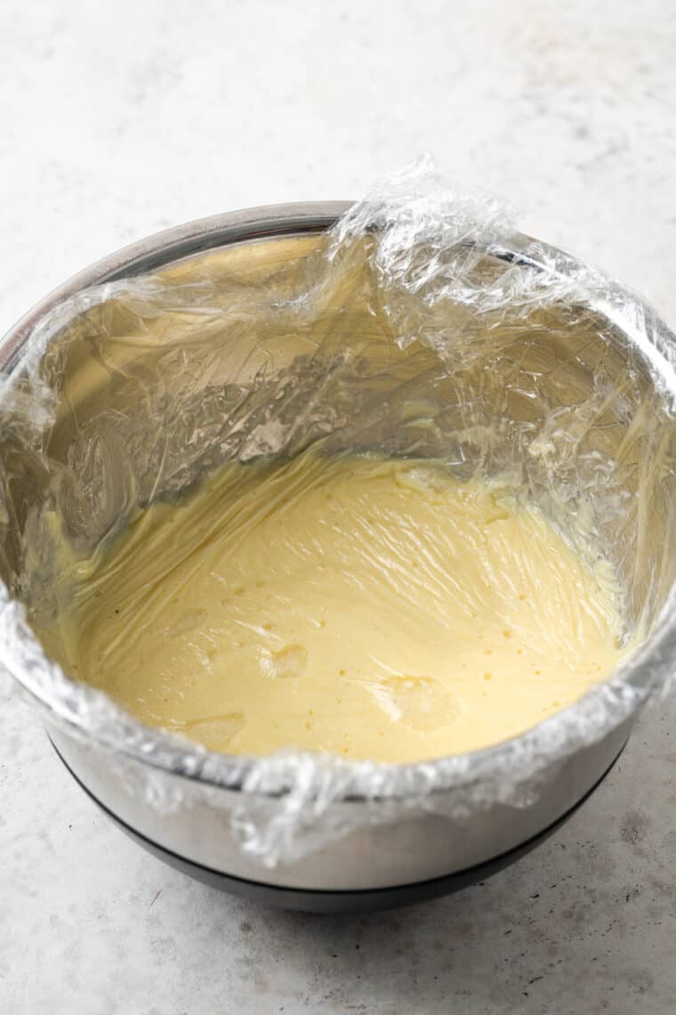 Pastry cream in a metal bowl is covered with plastic wrap.